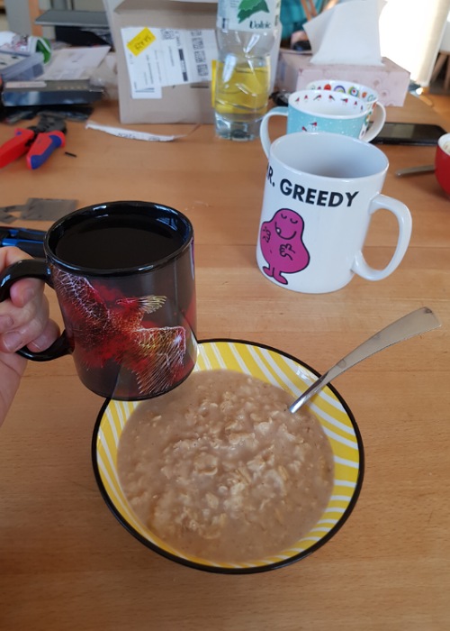 Mein Frühstückstisch: Du siehst eine große Schüssel mit Porridge. In der Hand halte ich eine schwarze Tasse mit Phönix-Motiv. 
